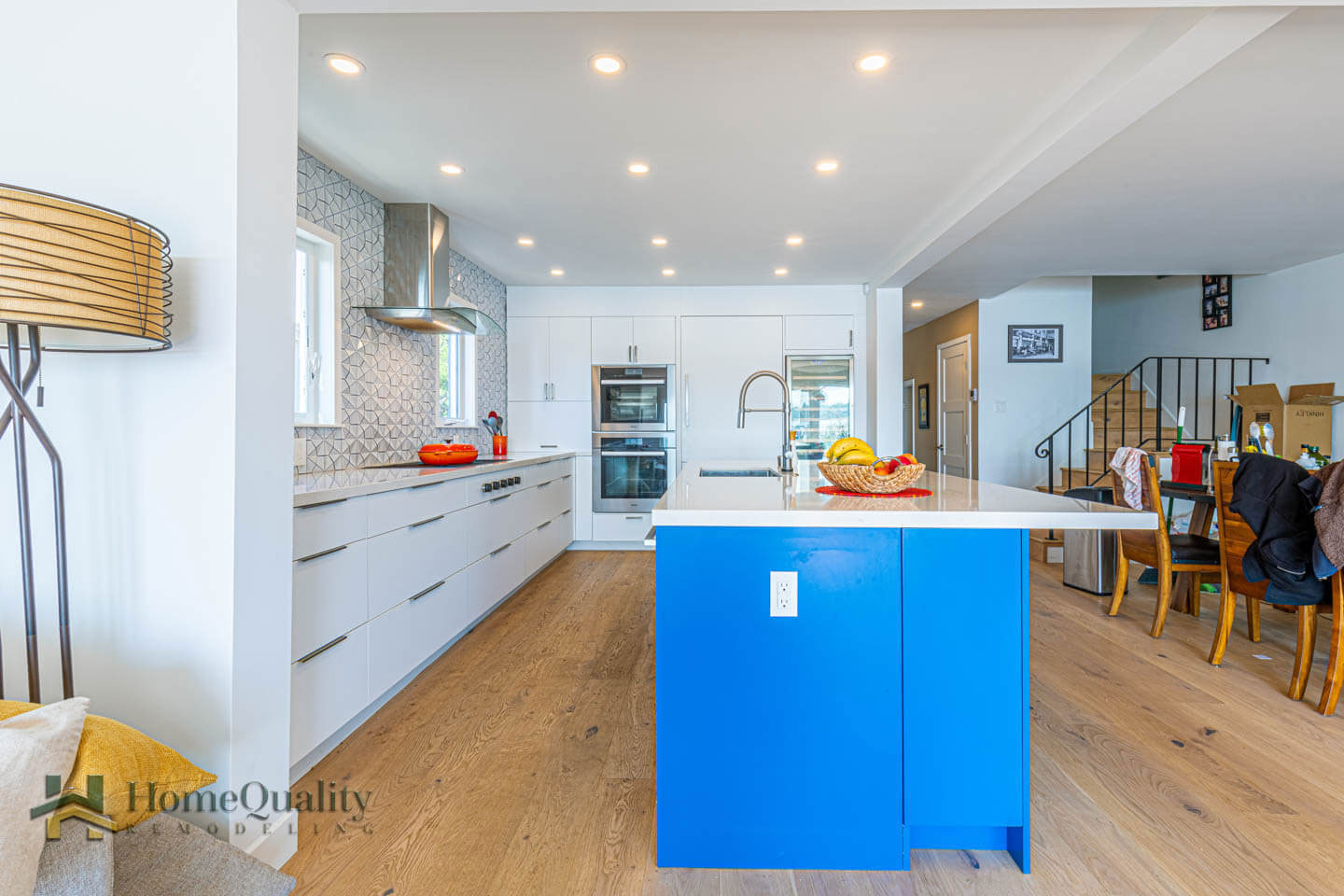 The image shows a modern kitchen interior with blue cabinets, white countertops, stainless steel appliances, hardwood flooring, and a central island with a blue countertop.