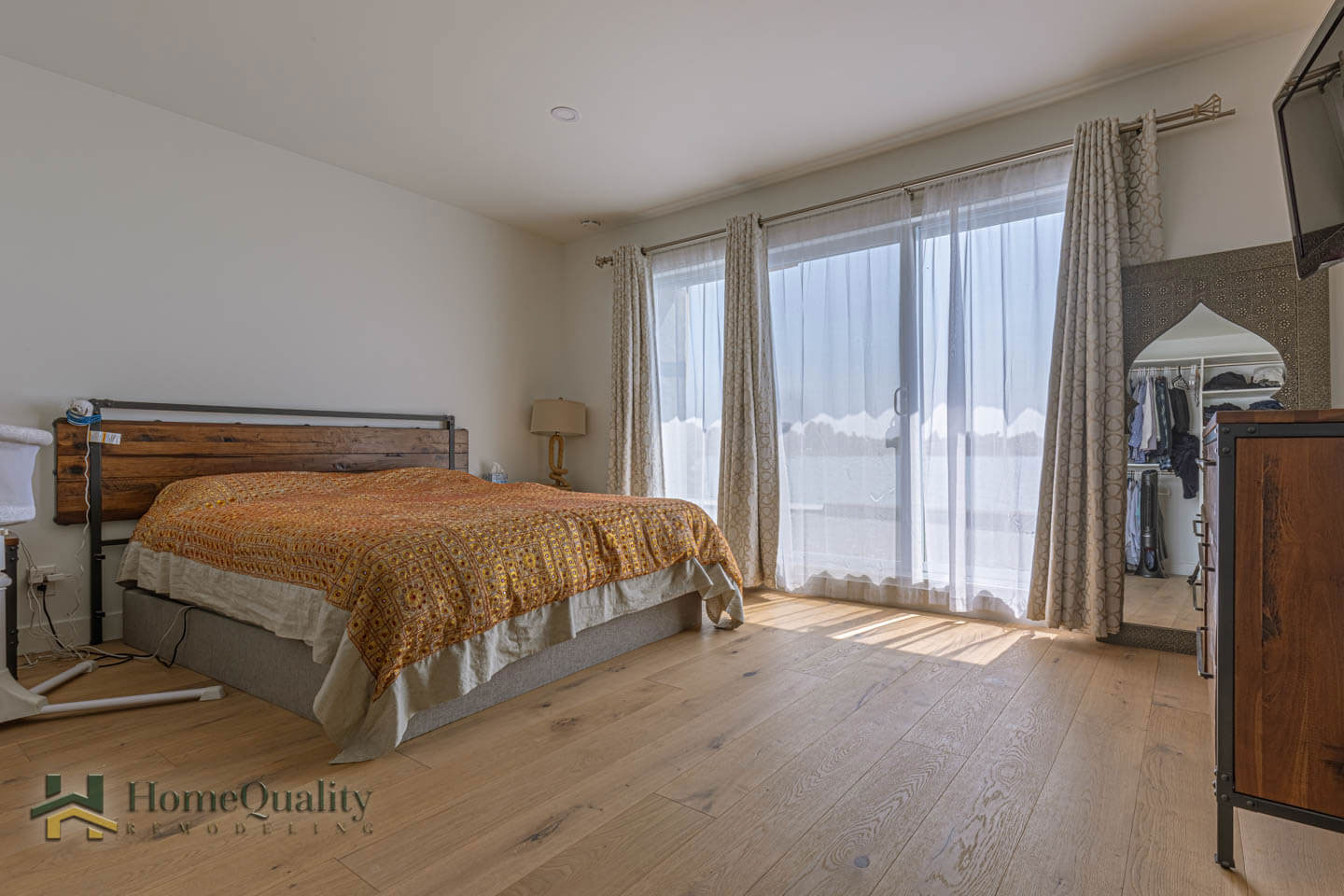 The image shows a neatly made hotel room with a large bed, wooden flooring, white walls, and curtains partially drawn over a window.
