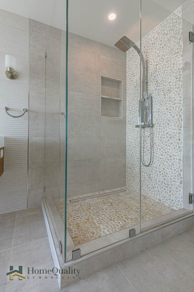 The image shows a modern bathroom with a walk-in shower featuring a glass door and a tile floor, inside which there is a handheld showerhead mounted on the wall above a built-in bench.
