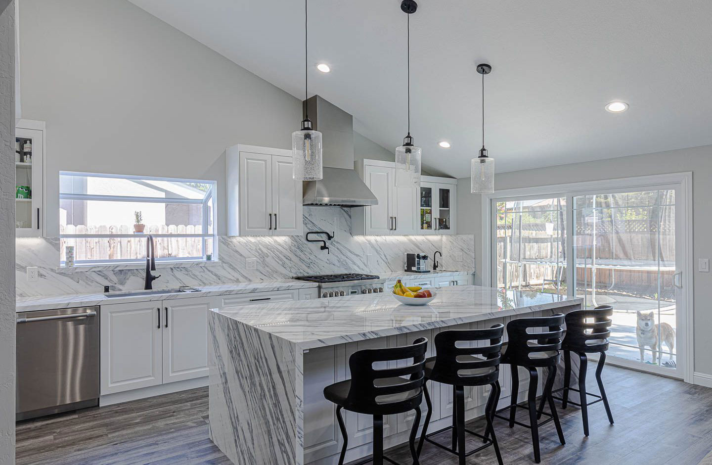 The image shows a modern kitchen interior with marble countertops, stainless steel appliances, a large island with barstools, pendant lighting, and a design that suggests a contemporary style.