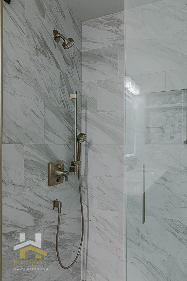 The image shows a modern bathroom with a marble-patterned wall, a glass shower enclosure, a chrome shower head, and a beige tiled floor.