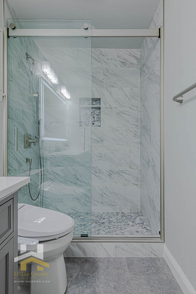 A modern bathroom with a glass shower door, marble walls, a white toilet, and a clean, minimalist aesthetic.