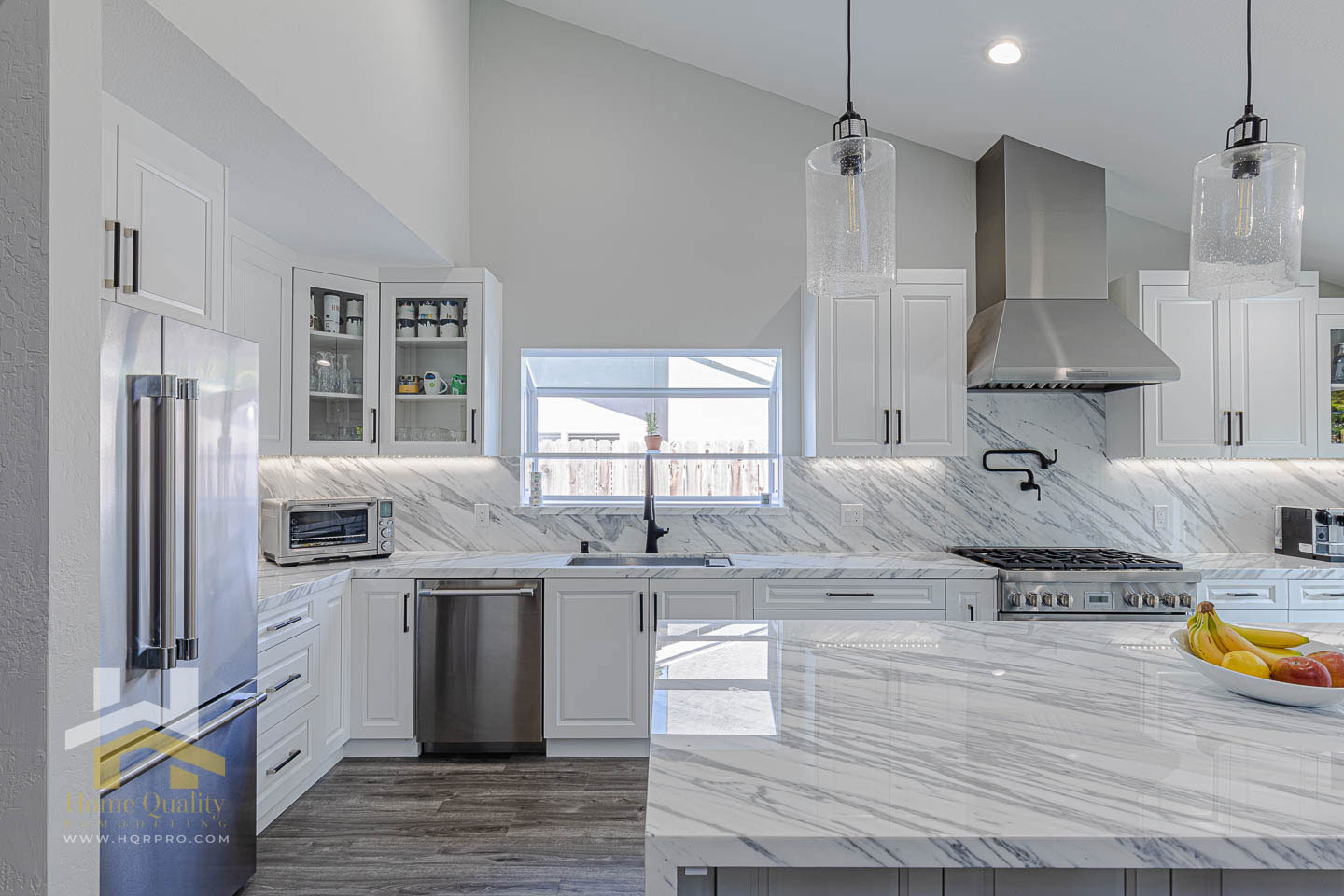 The image displays a modern kitchen with white cabinets, marble countertops, stainless steel appliances, and a large island featuring a built-in sink.