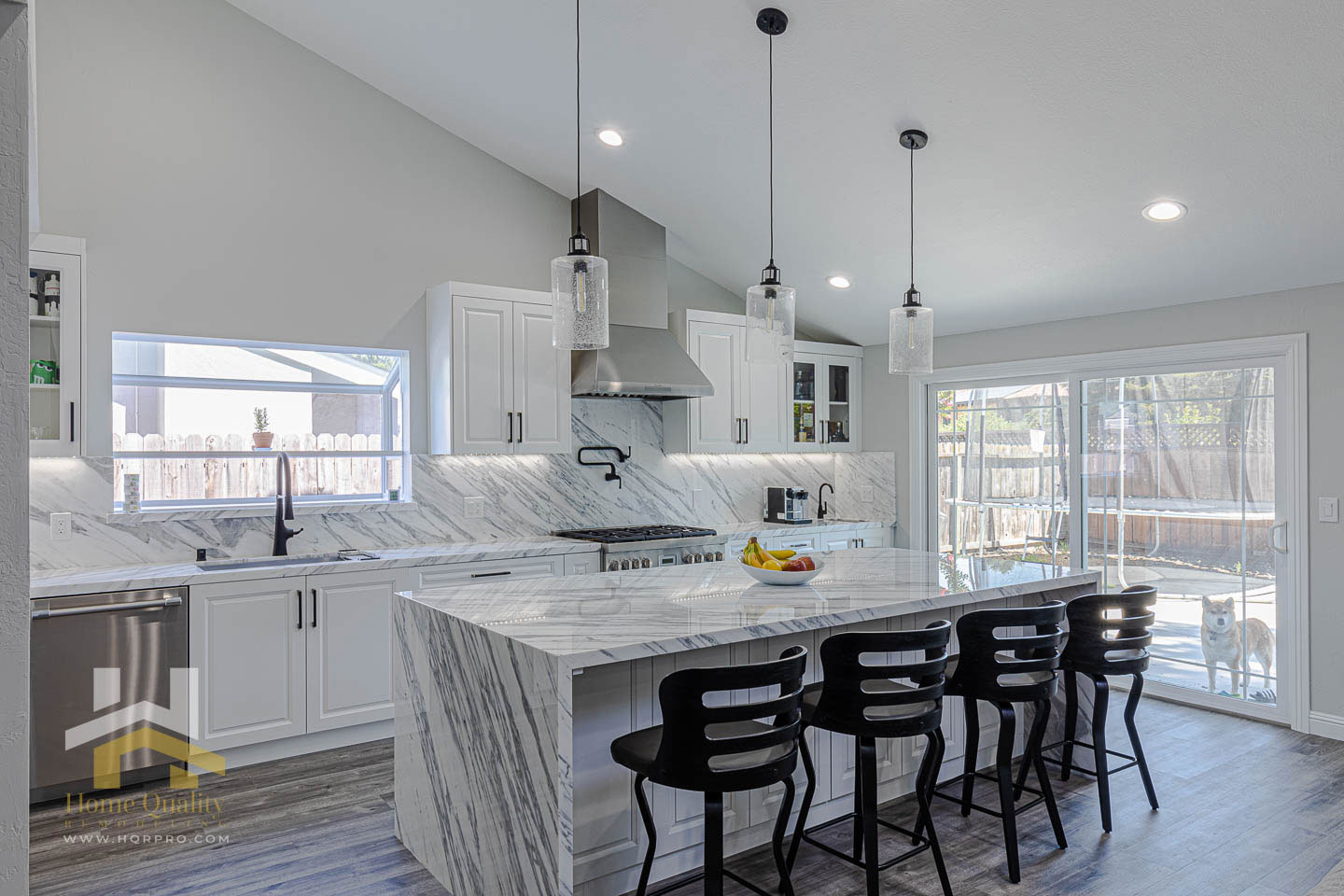 The image shows a modern kitchen with a large island in the center, featuring a marble countertop and stools, topped with a granite backsplash. The kitchen has white cabinets and stainless steel appliances, including a refrigerator and oven. A sink is installed under a window, and there are pendant lights hanging above the island. The flooring appears to be hardwood, and the ceiling has exposed ductwork.