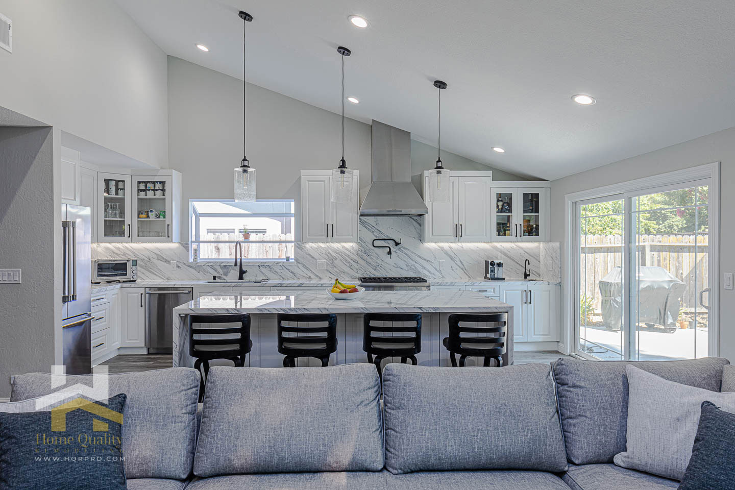 The image shows a modern kitchen with stainless steel appliances, granite countertops, and a center island, leading into an open-concept living area featuring a large sectional sofa and a contemporary gray color scheme.