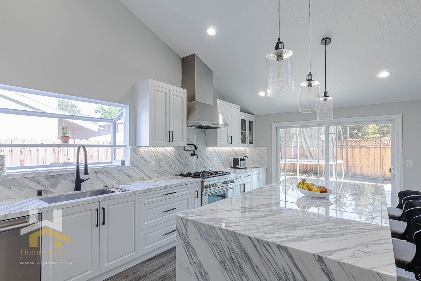 A modern kitchen with marble countertops and white cabinets, featuring a large island and stainless steel appliances.