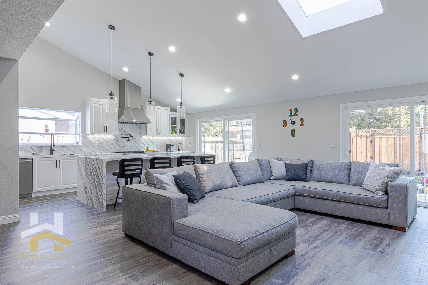 The image depicts a modern living room with a neutral color palette, featuring a large sectional sofa, a coffee table, a kitchen area with an island and countertop seating, a dining table, and a high ceiling with exposed ductwork and recessed lighting.