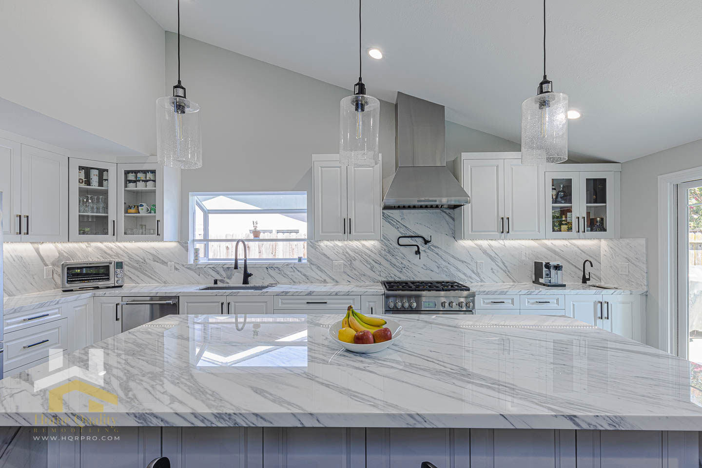 The image shows a modern kitchen with a large island featuring a marble countertop and a metallic finish, under-cabinet lighting, and a contemporary pendant light above the island.