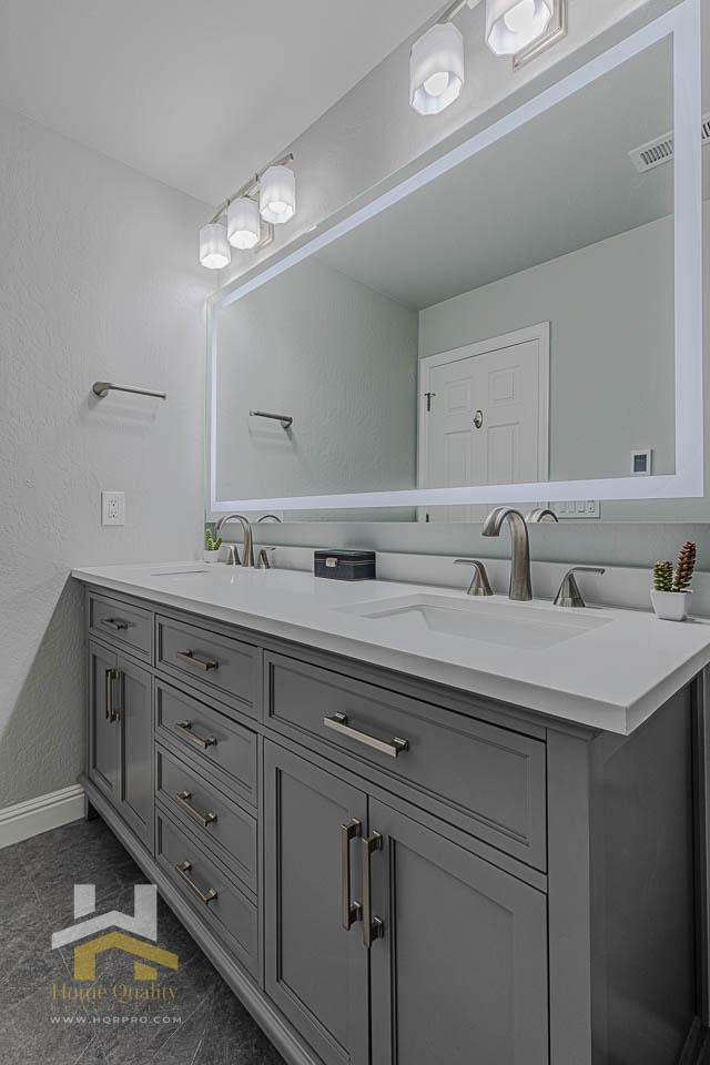 A modern bathroom with a large vanity mirror above a double sink countertop, featuring grey cabinets and white marble countertops.