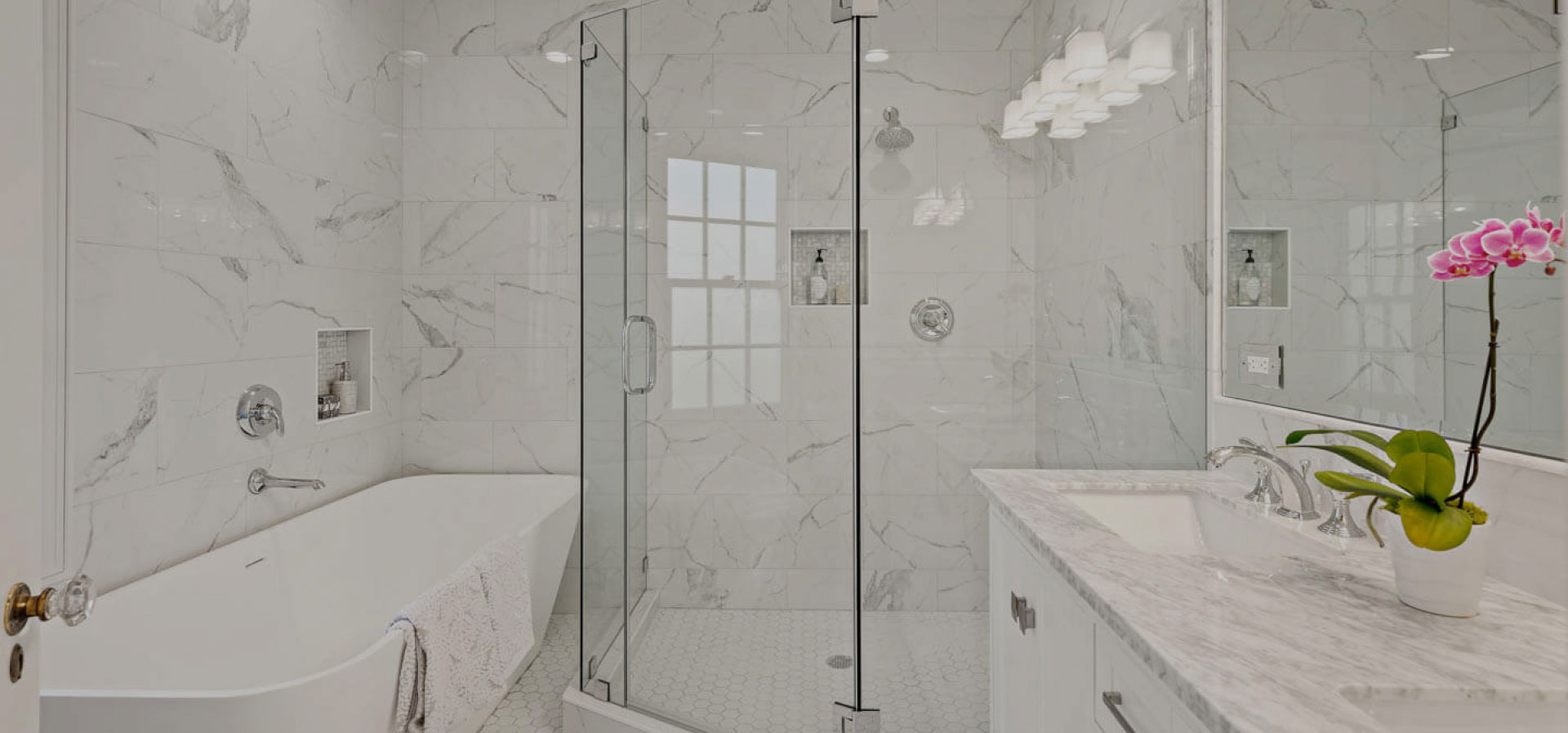 The image shows a modern bathroom interior with a glass-walled shower stall, marble walls and flooring, a white sink, and a large mirror above the vanity.
