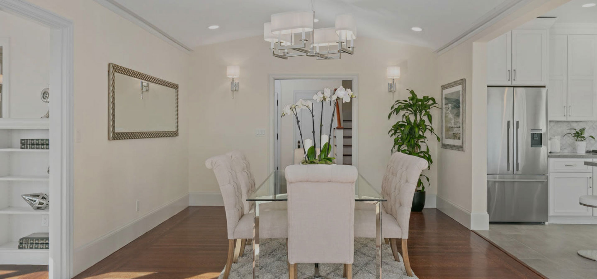 The image shows a modern living room with a kitchen area visible, featuring a contemporary design with neutral colors, white cabinets, stainless steel appliances, and a dining table set for four.
