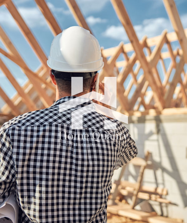 The image shows a construction worker wearing a hard hat standing on a building site with a blueprint in his hand, looking at a house frame under construction.