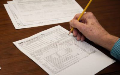 The image shows a person's hand holding a yellow pencil over a form with various checkboxes and lines for writing, placed on a wooden surface.