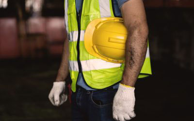 The image shows a person wearing a high-visibility vest, hard hat, and holding a clipboard, standing on a construction site with a backdrop of industrial equipment.