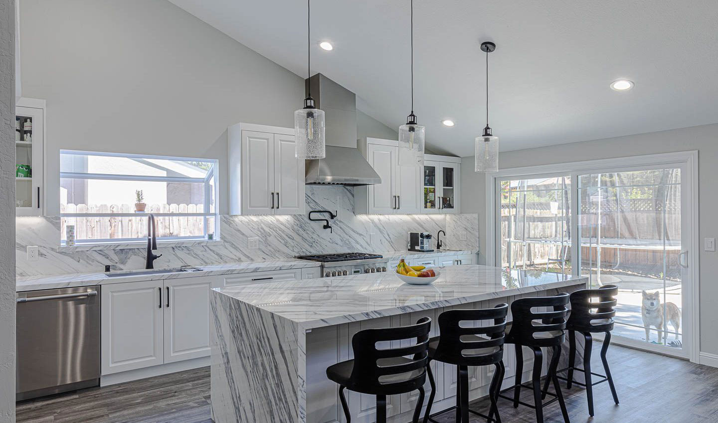 The image shows a modern kitchen with a large island at the center, featuring marble countertops, stainless steel appliances, pendant lighting above the island, and a dining area with barstools.
