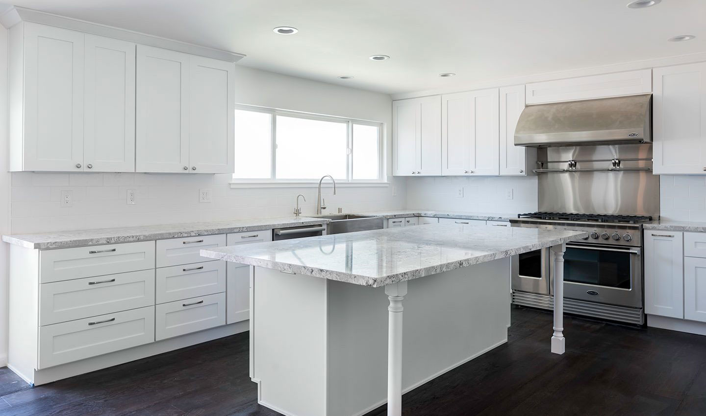 The image shows a modern kitchen interior with white cabinetry, a center island with a clear glass top, stainless steel appliances including an oven and refrigerator, and a large island countertop with a built-in sink.