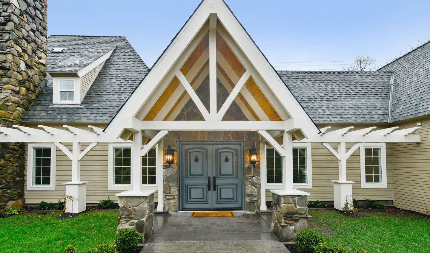 The image shows a two-story house with a prominent gable featuring a peaked roof design, accompanied by an attached garage with a matching white exterior and covered entrance. The house has a classic architectural style with visible wooden beams, suggesting a rustic or cabin-like aesthetic.